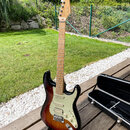 Elektrická kytara typu Stratocaster s tělem v provedení sunburst a světlým hnedým hmatníkem. Na hlavě je viditelné logo Fender. Kytara má krémový pic…