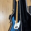 Modrá elektrická basová kytara s bílým pickguardem a hnedým hmatníkem, umístěná v černém tvrdém kufru. Na hlavě nástroje je viditelné logo Fender. Ky…