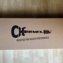 Hnědý papírový obal na hudební nástroj. Uprostřed je černé logo Charvel s nápisem MODIFIED FOR HIGHER PERFORMANCE a symbolem mostu. Na pravé straně j…