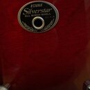Detail červeného lakovaného povrchu nástroje s oválným kovovým logem. Na logu je uveden nápis TAMA, Silverstar a ALL BIRCH SHELL spolu s číslem 07.9.…