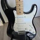 Černá elektrická kytara typu Stratocaster s bílým pickguardem a světlou hnedou hmatníkovou deskou. Nástroj má tři snímače, pětipositionní přepínač a…