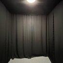 Fotografie zobrazuje malou, uzavřenou místnost s černými stěnami a stropem. V pozadí visí dlouhá černá látka, která slouží jako závěs. Podlaha je pot…