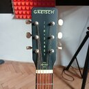 Černá hlava kytary s nápisem GRETSCH® v horní části. Uprostřed je svisle napsáno JIM + DANDY a v dolní části nápis STEEL REINFORCED NECK. Na hlavě js…