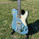 Modrá elektrická kytara s glitrovým povrchem a bílým pickguardem. Na mostku je vidět nápis Bigsby. Kytara má dva humbuckery a je umístěna na černém s…