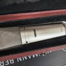 Stříbrný kondenzátorový mikrofon s kovovou síťovkou, umístěný v černé polstrované přepravce. Na těle mikrofonu je černý nápis WARM AUDIO a v zadní čá…