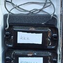 Dvě černé elektrické kytarové snímače v plastovém rámečku, umístěné na šedé pěnové výplasti v bílé krabičce. Snímače mají připevněné bílé papírové št…