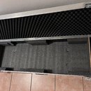 Otevřený hliníkový přepravní kufr s vnitřním polstrováním z černé pěny. Víko je vyloženo akustickou pěnou s vlnitým profilem, spodní část obsahuje vy…