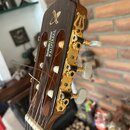 Hlava akustické kytary z hnědého dřeva se zlatými ladičkami a bílými knoflíky. Na zadní straně hlavy je zlaté logo ve tvaru stylizovaného písmena X a…
