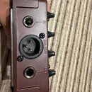 Detail boční strany hnědého elektronického zařízení. Jsou zde vidět tři konektory s nápisy IN, BAL OUT a OUT. Povrch je matný s mírnými známkami opot…