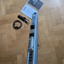 Stříbrný elektronický wind controller s nápisem AKAI EWI USB. K nástroji je přiložen černý napájecí adaptér s kabelem a dva papírové manuály s logem…