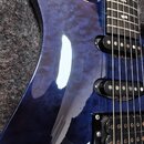 Detail elektrické kytary s modrým lakem a výraznou strukturou dřeva. Nástroj má tři černé humbuckery. Na povrchu laku jsou viditelné drobné škrábance…