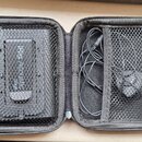 Otevřený černý přepravní pouzdro s vnitřními síťovými přihrádkami. V levé části je umístěno černé zařízení s nápisem RIG ACOUSTIC Stage. V pravé část…