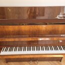 Hnědé dřevěné piano s lakovaným povrchem. Na čelní straně nad klávesami je zlatý nápis PETROF. V otevřeném horním víku jsou vidět struny a v pravé čá…