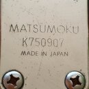 Detail kovového šiltiku s nápisy MATSUMOKU, K750907 a MADE IN JAPAN. Šiltík je připevněn čtyřmi křížovými šrouby a vykazuje drobné škrábance a známky…