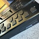 Černý elektronický hudební přístroj s řadou gumových tlačítek. Na horním panelu je vidět nápis ASR a označení Transport s doprovodnými popisky. Povrc…