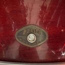 Detail kovového oválného štítku na červeném lakovaném povrchu bubnu. Na štítku je nápis TAMA, ROCKSTAR a MADE IN TAIWAN spolu s číslem 323805. Povrch…