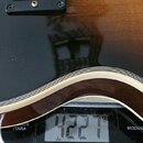 Detail těla kytary s tmavým, lesklým povrchem a bílým lemováním. Na kytáře je vidět přepínač s nápisy TREBLE a RHYTHM. Nástroj leží na digitální váze…