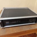 Černý výkonový zesilovač QSC AUDIO BMX 1850HD PROFESSIONAL POWER AMPLIFIER umístěný v transportním flightcasu s hliníkovými hranami. Na předním panel…