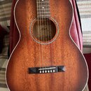 Akustická kytara s hnědým dřevěným povrchem a bílým lemováním. Uvnitř otvoru pro zvuk je vidět štítek s nápisem Cort Acoustic. Kytara má tmavý hmatní…