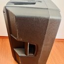 Černý plastový reproduktor s hranatým tvarem a integrovanými úchyty na horní a boční straně. Povrch je matný s viditelnými odpadenými částicemi prach…