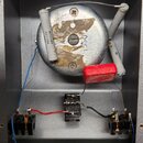 Interiér šedého kovového šasi s elektronickými komponenty. Uprostřed je kulatý kovový prvek s nápisem 1360, vedle něj se nachází červený obdélníkový…