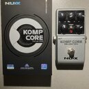 Na fotografii je stříbrný efektový pedál s černými ovládacími knoflíky a přepínačem. Na pedálu jsou nápisy KOMP CORE DELUXE MKII a logo NUX. Vedle pe…