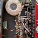 Fotografie zachycuje vnitřek elektronického zařízení, pravděpodobně zesilovače. Je zde vidět velký toroidní transformátor zabalený v průhlednou fólii…