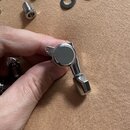 Fotografie zobrazuje chromované ladicí mechanismy na kytaru. V popředí drží ruka jeden kompletní ladicí kolík s knoflíkem, v pozadí jsou rozložené da…