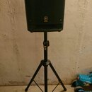 Černý reproduktor umístěný na černém stativu se třemi nohama. Na přední straně reproduktoru je zlaté logo a nápis YAMAHA. Stav povrchů vypadá čistě,…