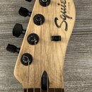 Fotografie zachycuje hlavu elektrické kytary z přírodního světlého dřeva s černými ladičkami. Na hlavě je čitelně napsáno logo Squier by Fender a náp…