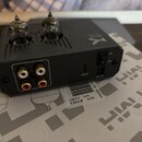Černý elektronický přístroj s dvěma vakuumovými elektrony v horní části. Na zadní straně jsou čtyři RCA konektory s nápisem AUX, USB port označený ja…