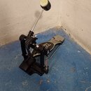 Černý bicí pedál s řetízkovým pohonem a kovovou šlapkou. Na šlapce je viditelný nápis TAMA. Beater má na konci bílou plfeltovou hlavici. Nástroj vyka…