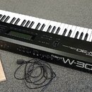 Černý klavierní syntetizátor s bílými a černými klávesami. Na předním panelu jsou nápisy Roland W-30 a Roland MUSIC WORKSTATION. Vedle nástroje je po…