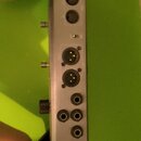 Stříbrný kovový panel s řadou různých konektorů a vstupů. Jsou zde vidět dva XLR konektory, několik jackových vstupů a napájecí zásuvka. Na panelu ne…