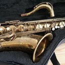 Zlatý saxofon v černém pouzdře. Na povrchu zvonu jsou viditelné ornamentální vzory a známky opotřebení v podobě drobných škrábanců a patiny. K nástro…