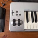 Stříbrná elektronická klaviatura s bílými a černými klávesy. Na ovládacím panelu jsou nápisy VOLUME, ADVANCED FUNCTIONS, OCTAVE, PITCH BEND a MODULAT…