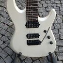 Bílá elektrická kytara s hnědým hmatníkem a třemi černými humbuckery. Nástroj je umístěn na černém stojanu na kamenné dlažbě. Na těle jsou vidět dva…