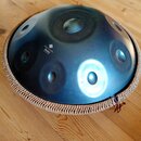 Kovový perkusní nástroj v podobě modrého polokoule s několika otvory. Na povrchu je bílý nápis Melody Handpan Sela handpan. Okraj nástroje je opleten…