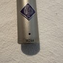 Detail stříbrného kovového těla mikrofonu. Na povrchu je modré kosoctytý logo s nápisem NEUMANN a stylizovaným symbolem. V dolní části je vyrazeno sé…