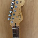 Fotografie zachycuje hlavu a hриф elektrické kytary z přírodního dřeva s hnědým hmatníkem. Na hlavě je viditelné logo Fender a nápis STRATOCASTER®. V…