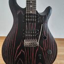 Elektrická kytara s černým matným povrchem a červenými prouhy v imitaci zrn dřeva. Nástroj má dva humbuckery, jeden ovládací přepínač a dva otočné kn…