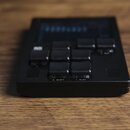 Černý elektronický přístroj s displejem a několika ovládacími tlačítky, položený na dřevěném povrchu. Na spodní hraně jsou viditelné nápisy SHIFT a P…