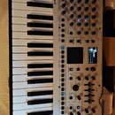 Bílý syntetizátor s klaviatura značky Roland, model GAIA 2. Přístroj má numerous ovládací prvky v podobě posuvníků, otočných knoflíků a tlačítek, vče…