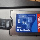 Na fotografii jsou dva adaptéry značky SanDisk. Větší černý adaptér má modře a červeně barevnou nálepku s nápisy SanDisk a 6-in-1 PC Card Adapter. Ve…