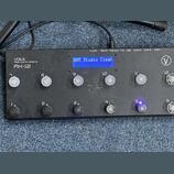 Černý MIDI kontrolér s nápisy VOES Midi Controllers a MX-12. Zařízení má modrý displej s textem 005 Studio Clean a dvanáct tlačítek označených čísly…