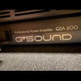 Detailní záběr na přední panel černého zesilovače. Je na něm bílý nápis Professional Power Amplifier QSA 600 a pod ním logo QSOUND. Vlevo je vidět vy…