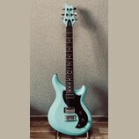 Světle modrá elektrická kytara s černým pickguardem a hnědým hmatníkem. Na hlavě je bílé logo "Cort" a nápis "Veta". Kytara má dva humbuckery, jeden…