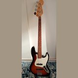 Elektrická basová kytara s tělem v provedení sunburst a bílým pickguardem. Na hlavě hředu je viditelné logo Fender. Nástroj má dřevěný hmatník a čtyř…