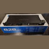 Černý rackový audio interface značky MOTU, který leží ve své původní krabici. Na předním panelu jsou vidět dva kombinované konektory, ovládací prvky…