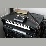 Černý rack s dvěma vloženými zařízeními. Horní jednotka má stříbrný přední panel s řadou bílých oválných knoflíků. Na vrchu racku leží černý box s bí…